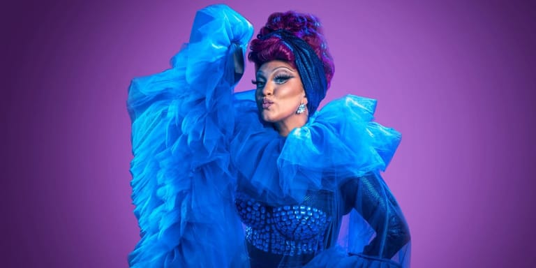 Performer in dramatic blue ruffled costume and sunglasses against vibrant purple backdrop.