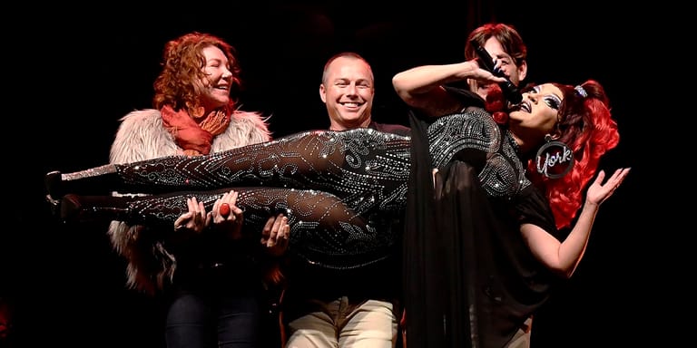 Four performers playfully pose with a large fake fish during a comedic stage production.