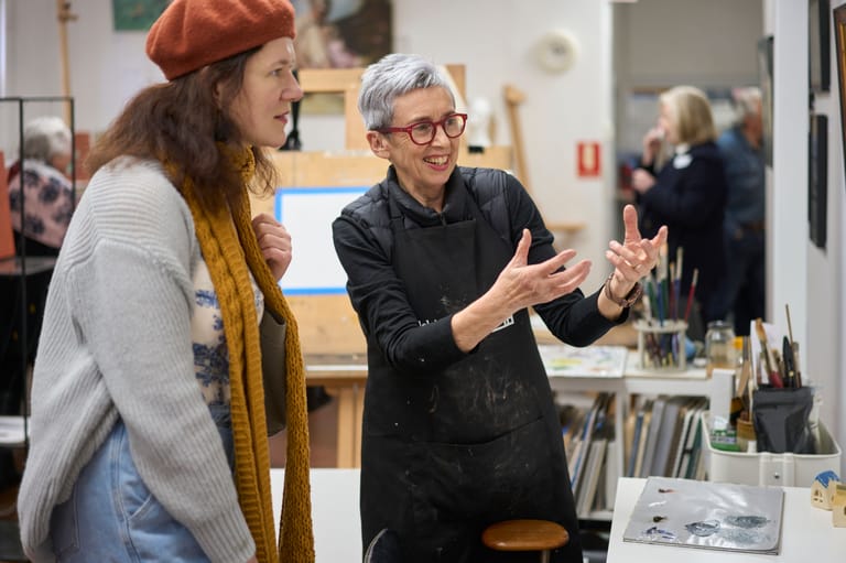Two people converse in an art studio or workshop space, with art supplies and other visitors visible in the background.
