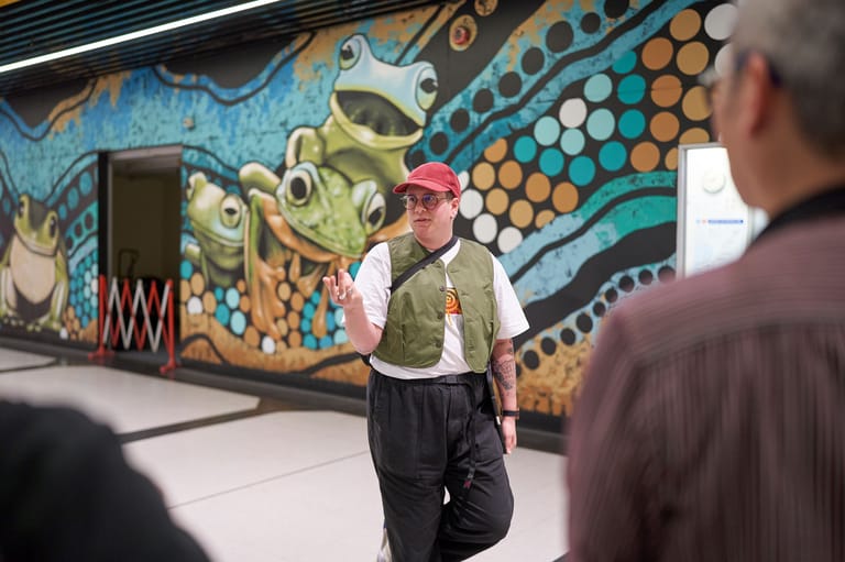 A man in a pink cap and olive vest stands before a vibrant blue and gold mural featuring stylised frog designs.