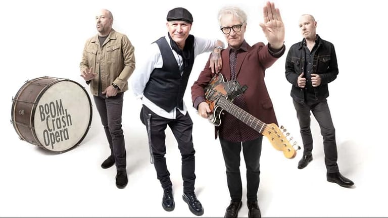 Five band members pose against white background, with drummer on left and guitarist centre-front waving to camera.
