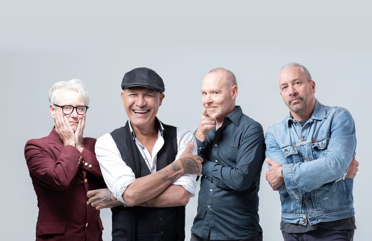 Four men in casual and formal attire stand against a grey background in a relaxed group pose.