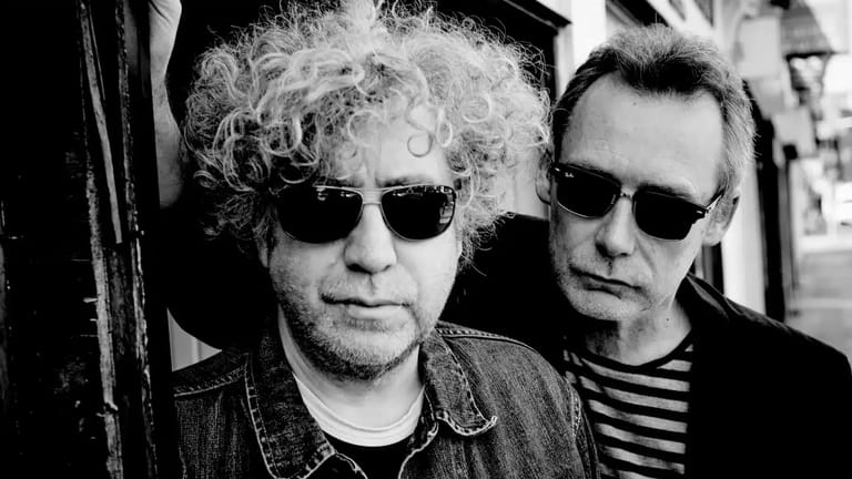 Two men wearing sunglasses pose together in black and white, one with voluminous curly hair.
