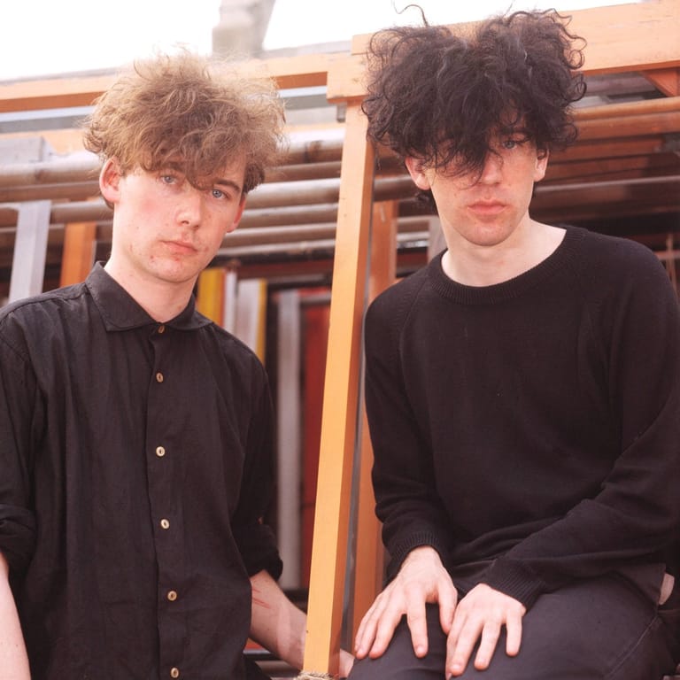 Two young men with tousled dark hair sit against wooden beams in an indoor setting.