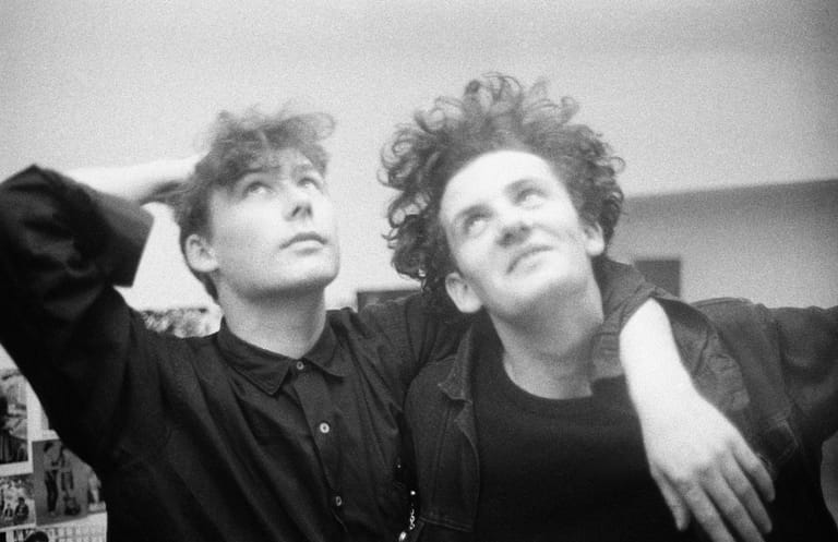 Two people with tousled dark hair sit together in black and white, looking upward thoughtfully during an interview setting.