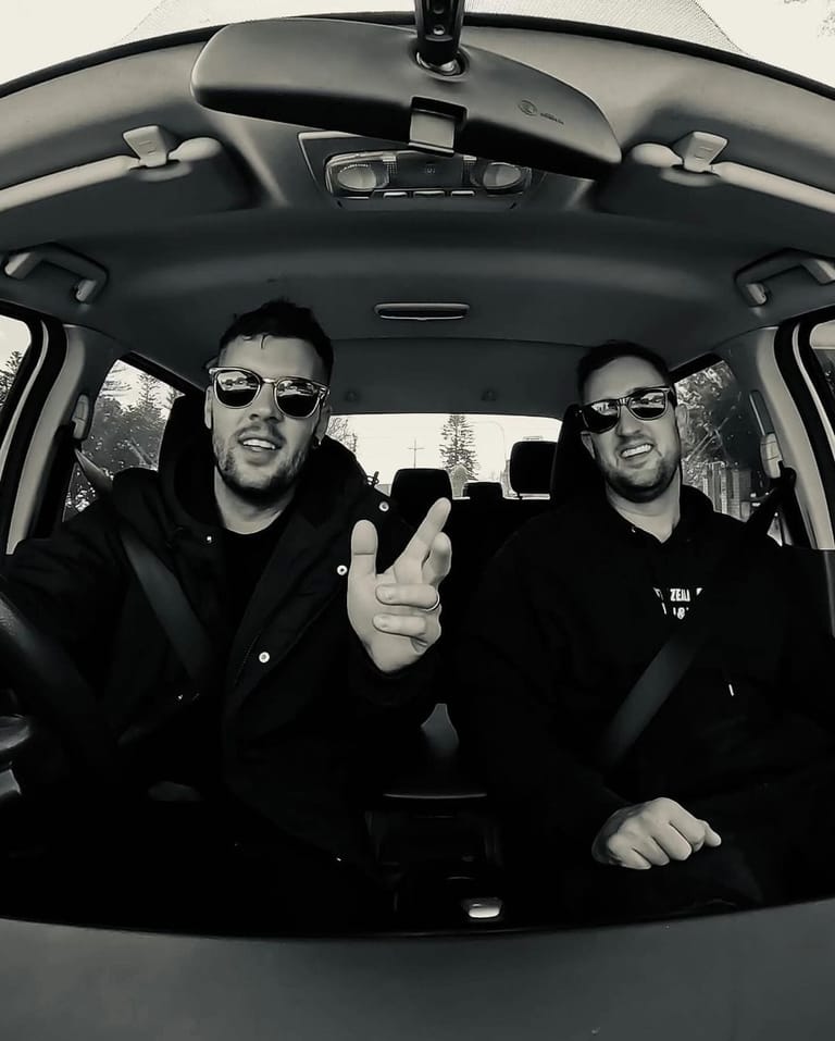Two people wearing sunglasses sit in a car interior, one gesturing toward the camera in a black and white photograph.