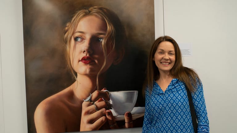 Woman in blue patterned jacket stands beside a portrait painting of a figure holding a cup.