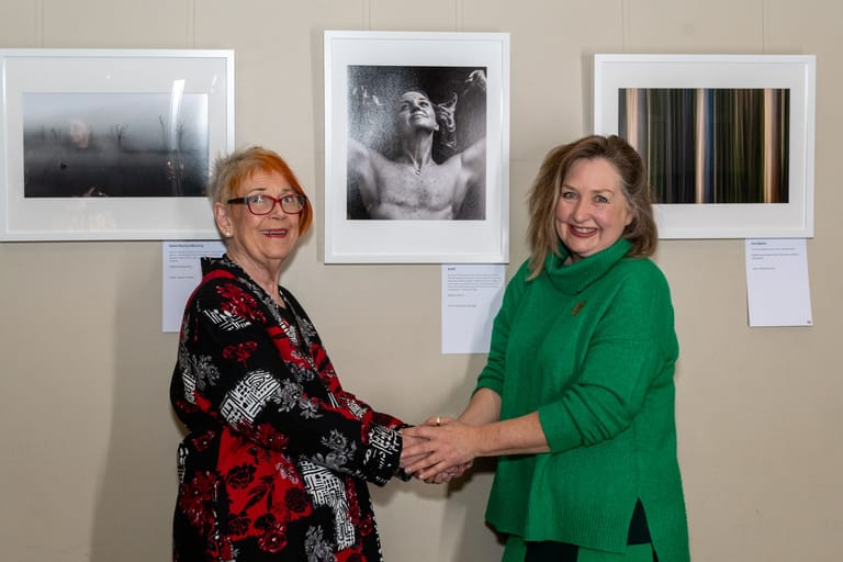 Two women shake hands in front of framed photographs displayed on a gallery wall.