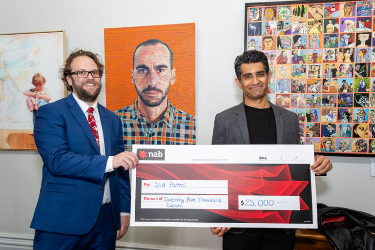 Two men in formal attire present a large cheque in an art gallery surrounded by colourful portraits and artworks.