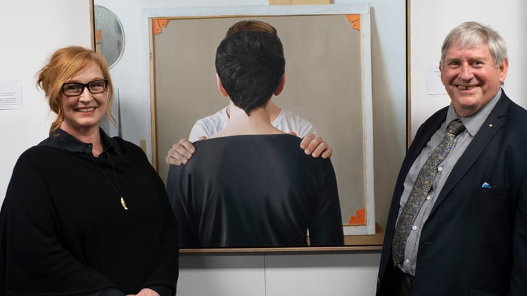 Two people flank a framed artwork featuring a figure viewed from behind against a neutral background.