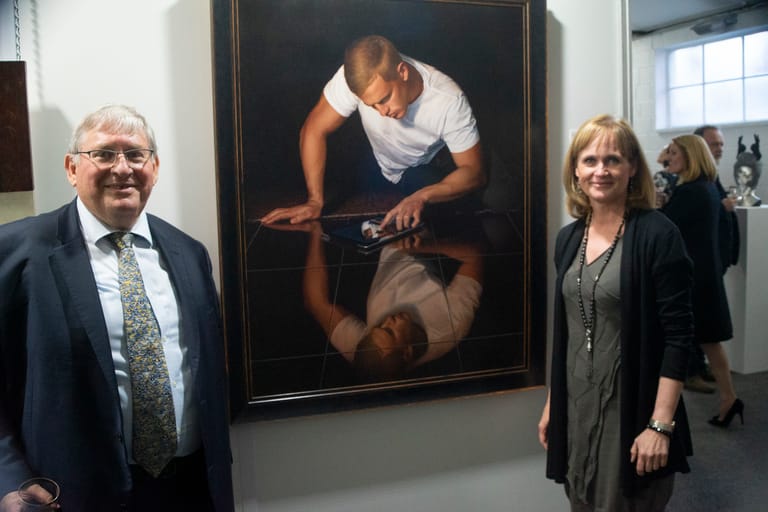 Two people stand beside a large portrait painting of a man in a white shirt against a dark background in a gallery space.