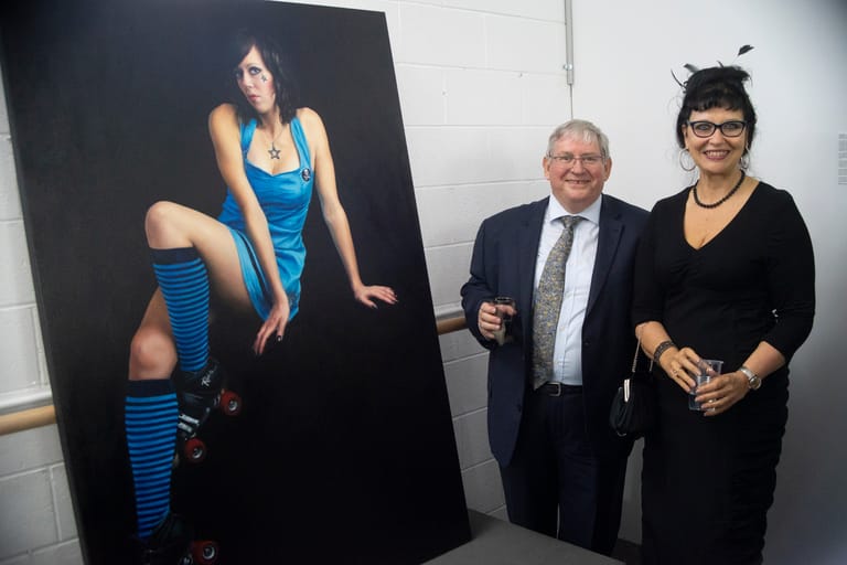Two people stand beside a large portrait of a woman in a blue sequined outfit against a white gallery wall.