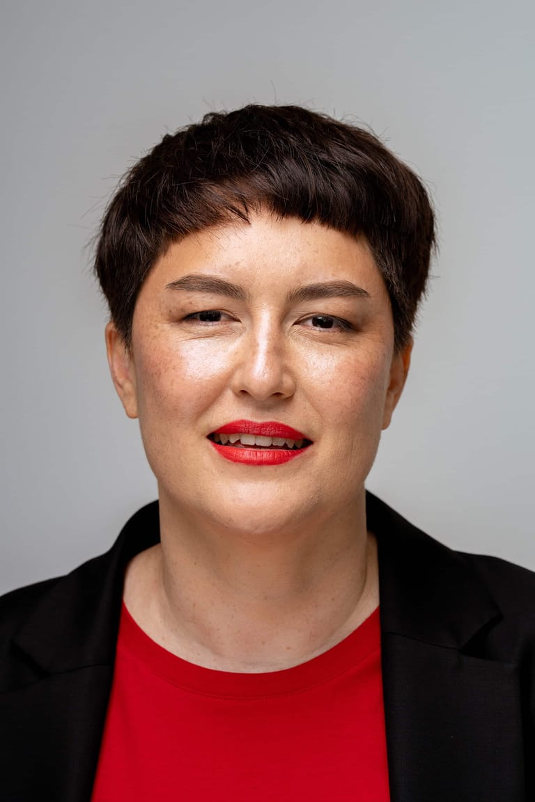 Person with short dark hair and red lipstick wearing a red shirt and black blazer against a grey background.