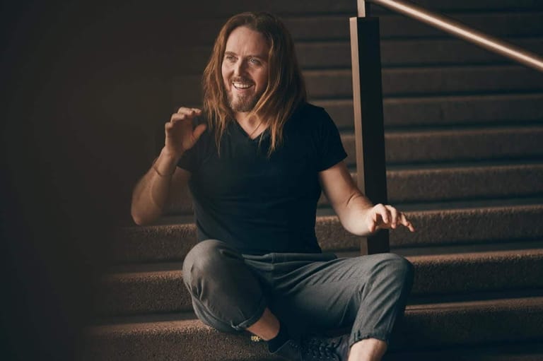 Man with long hair crouching on wooden stairs, smiling at camera in warm stage lighting.