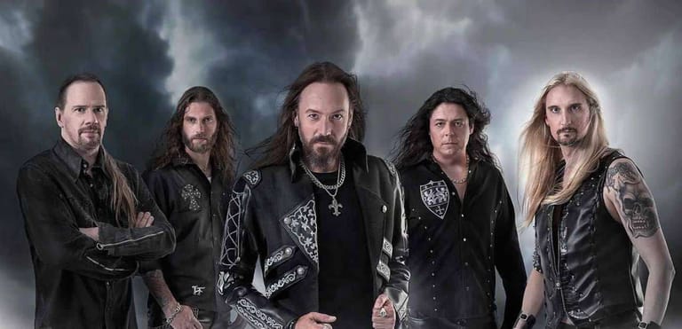 Five musicians in black leather and metal jewellery pose against a dramatic stormy sky backdrop.