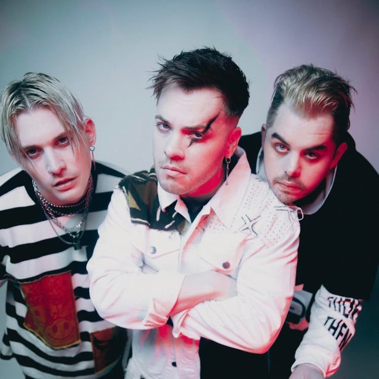 Three band members in striped and plain shirts face the camera with pink and red stage lighting casting shadows across their faces.