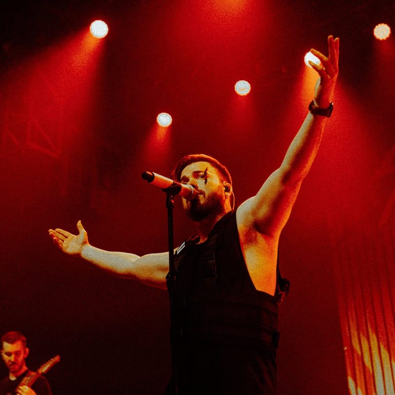 Singer at microphone with arm raised, bathed in red stage lighting and spotlights.