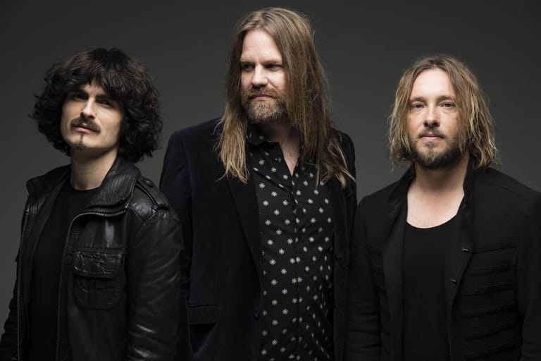 Three men with long hair and facial hair stand against a grey background in a studio portrait.