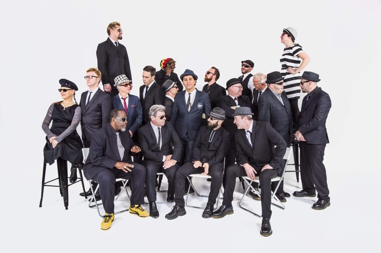 Large ensemble of performers in black suits and hats posed together against a white background.