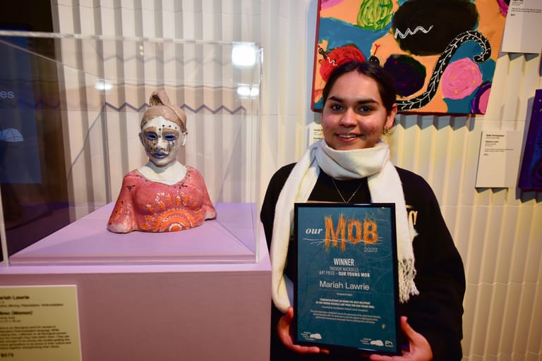 Person holding an award certificate stands beside a display case containing a decorative masked bust in an art gallery setting.