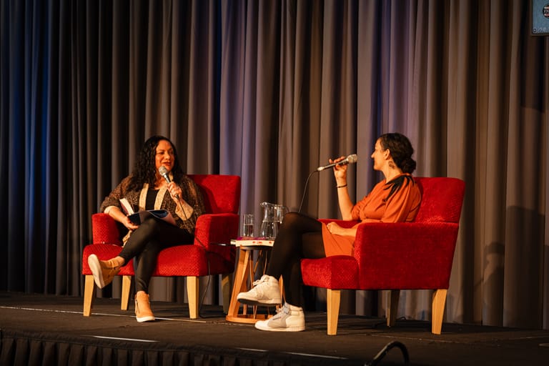 Two women seated in red chairs on stage during an intimate conversation or interview under warm stage lighting.