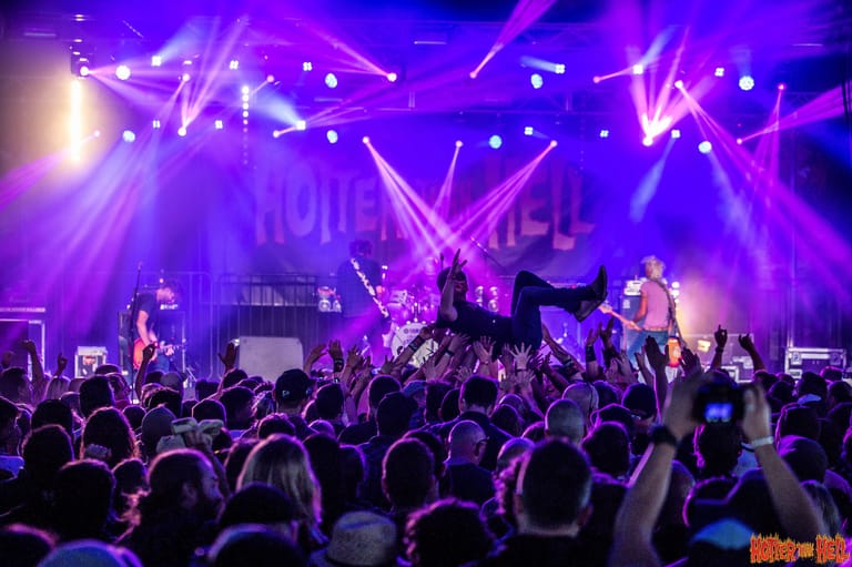 Crowd of concert-goers with raised arms facing a stage lit by purple and white spotlights during a live music performance.