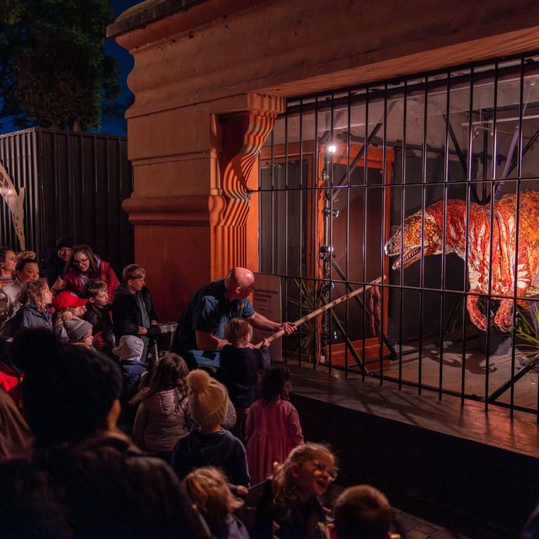 Audience members gather before a theatrical performance featuring a caged figure illuminated by warm orange lighting.