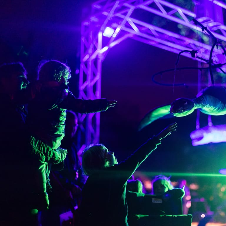 Performer in neon green clothing gestures expressively on stage beneath purple lighting rigs during a live show.