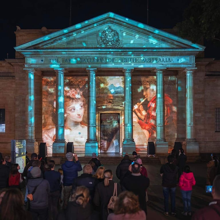 Classical building facade illuminated in turquoise light with projected artworks displayed on windows, spectators gathered in front at night.