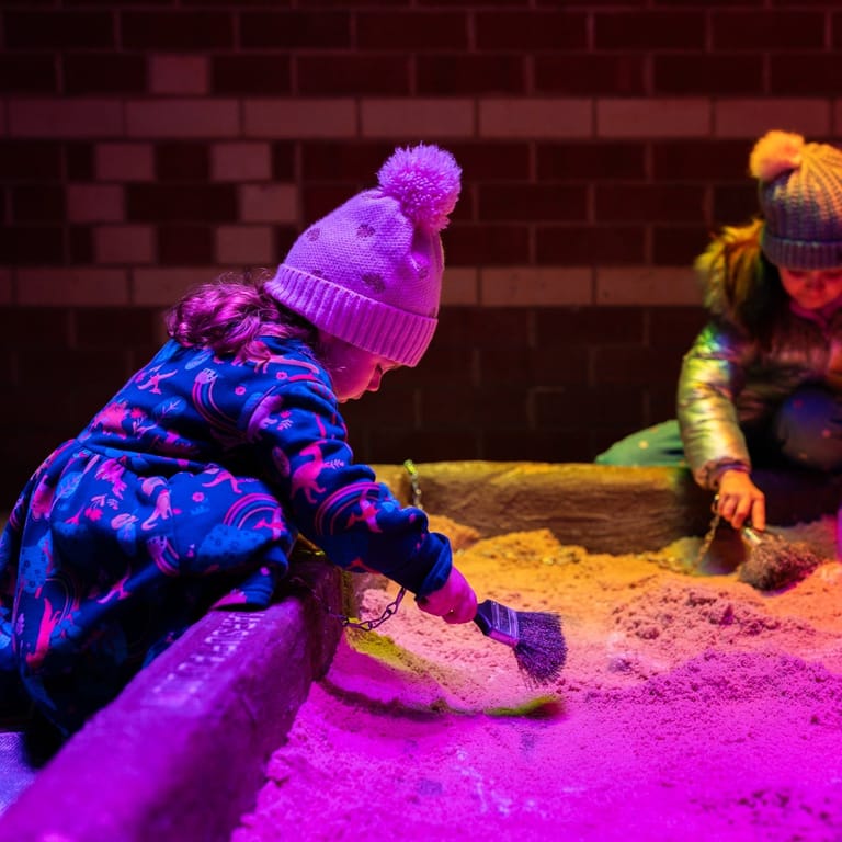 Two children in winter clothing crawl through sand under vibrant pink and purple neon lighting.