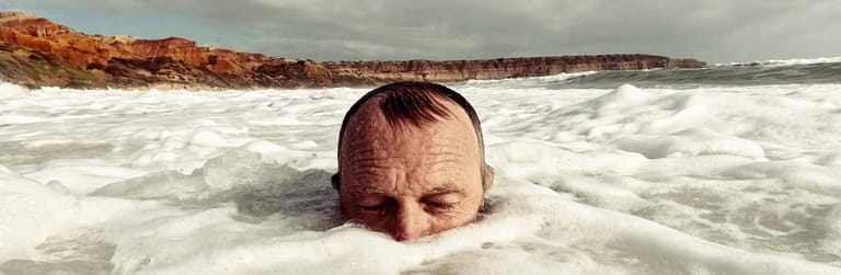 Man's head submerged in foamy ocean waves at a sandy beach beneath overcast sky.