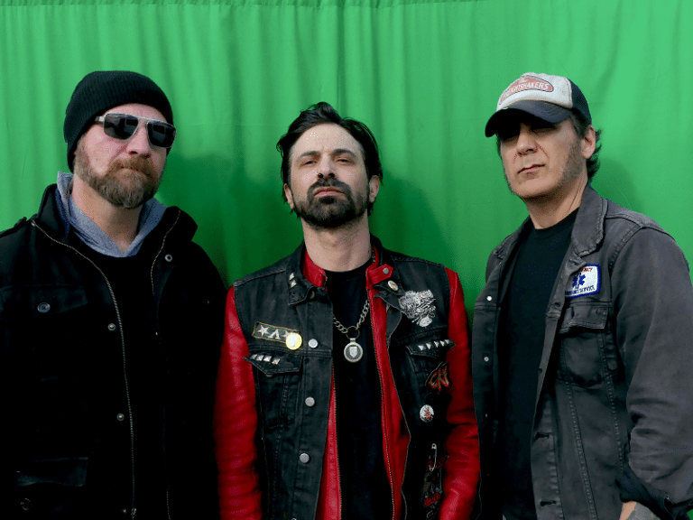 Three musicians wearing sunglasses and caps stand against a bright green backdrop.