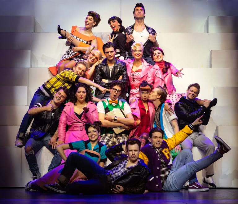 Large ensemble cast in vibrant costumes poses dynamically on stage in a pyramidal formation.