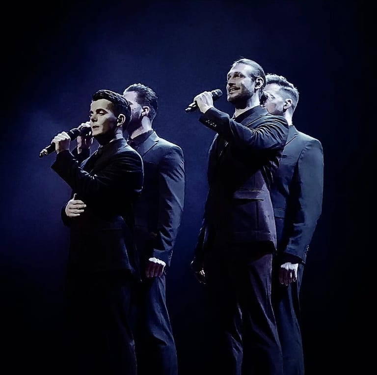 Four male singers in dark suits performing together on stage under blue spotlight lighting.