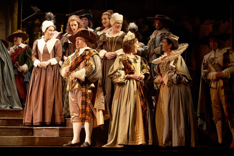 Ensemble of performers in elaborate Renaissance-style costumes and gold fabric on a dimly lit stage.