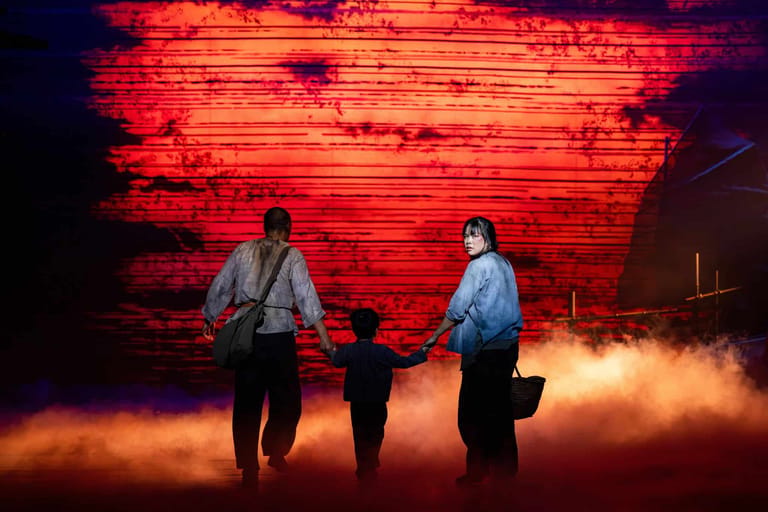 Three figures stand silhouetted against a glowing red neon wall with dramatic orange fog billowing across the stage.