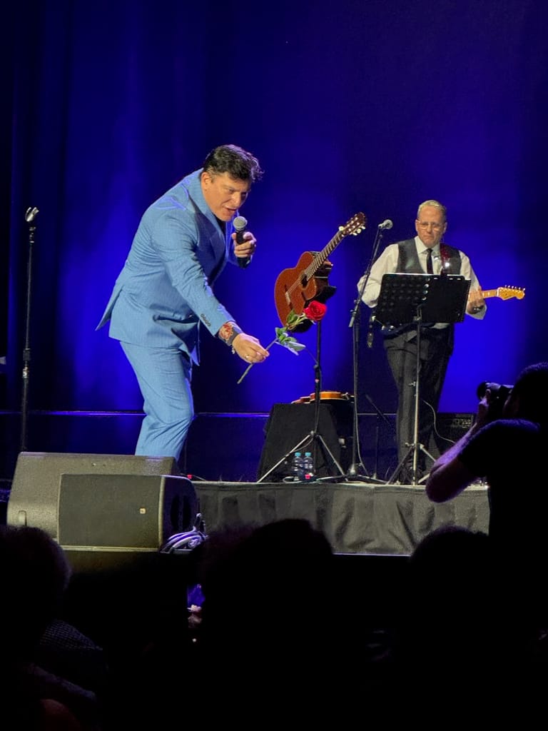 Singer in light blue suit performs with microphone as guitarist and keyboardist accompany on stage beneath blue lighting.
