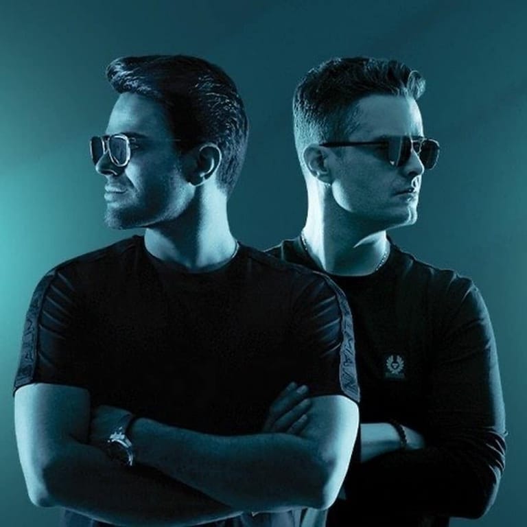 Two men wearing sunglasses and dark shirts stand back-to-back against a teal-blue lit background.