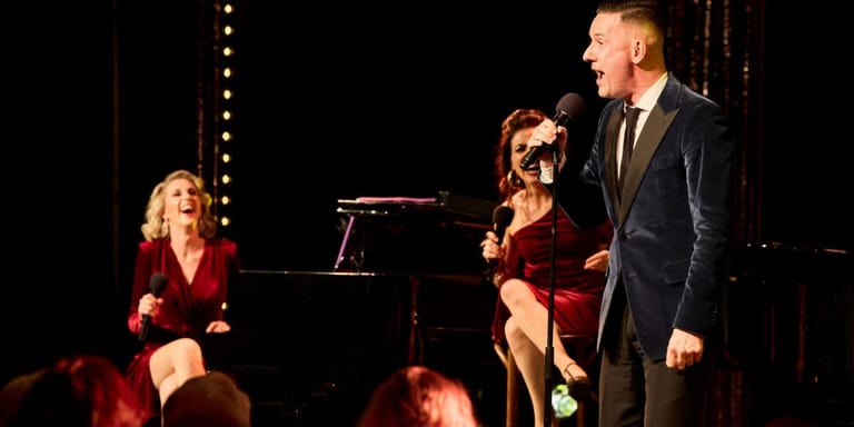 Three performers in formal attire on a dimly lit stage with a piano and decorative lights behind them.