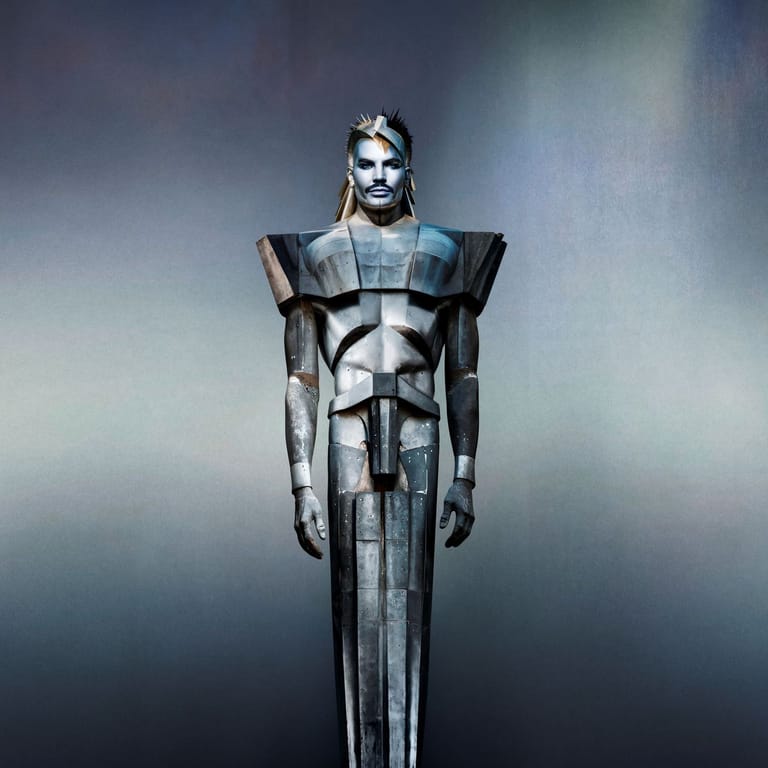 Figure in bold geometric armour and pale makeup stands against a grey, atmospheric background.