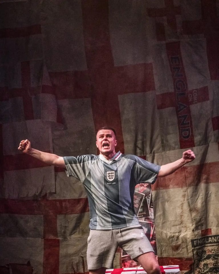 Performer in England football kit with arms outstretched against a large Union Jack backdrop on stage.