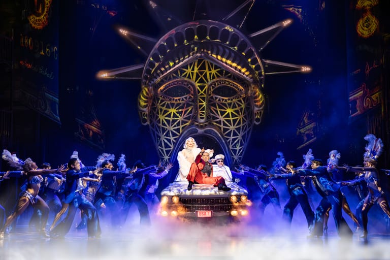 Performer in elaborate costume seated centre stage beneath ornate golden mask projection, surrounded by ensemble cast in blue lighting and atmospheric fog.