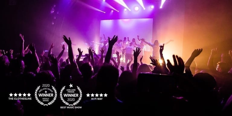 Silhouetted crowd with raised hands dancing under purple and amber stage lighting at a concert venue.