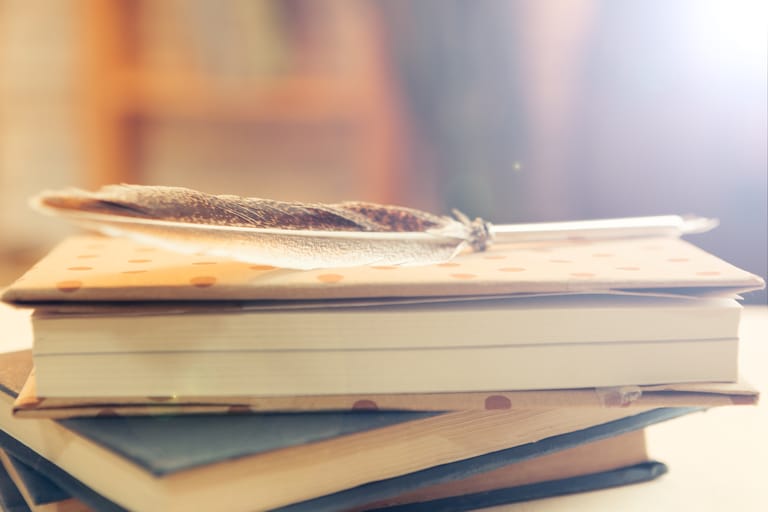 A feather rests on a stack of closed books in warm, soft lighting.