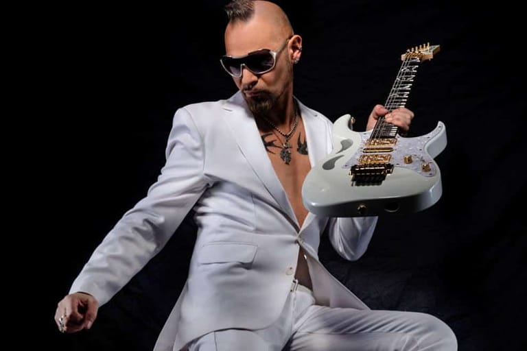 Musician in white suit and sunglasses holding white electric guitar against black background.