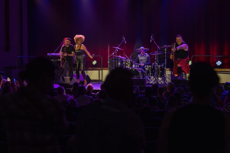 Band performing on stage under purple and red stage lighting with audience silhouettes in foreground.