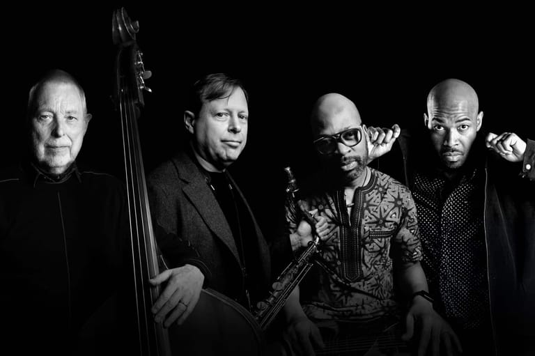 Four musicians in black and patterned clothing posed against a dark background, one holding a double bass.