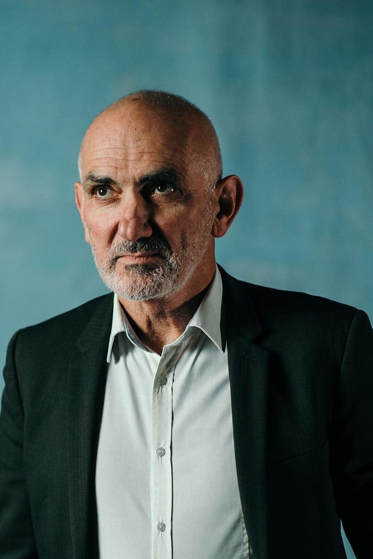 Man with grey beard wearing dark blazer and white shirt against teal blue background.