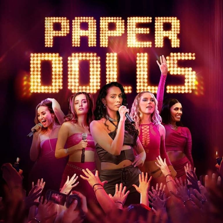 Five performers in pink and purple outfits stand against a glowing "Paper Dolls" marquee with raised hands below.