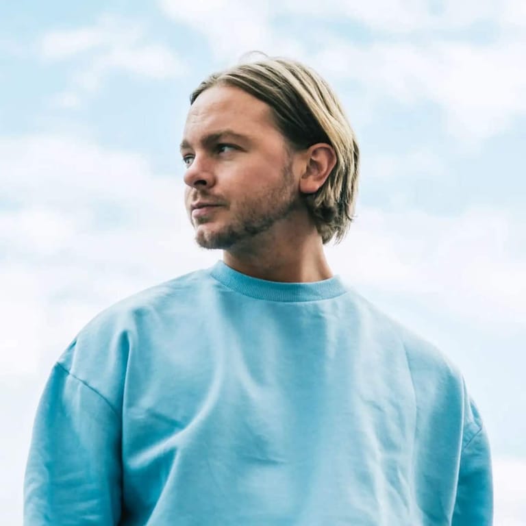 Man with shoulder-length blonde hair wearing a light blue t-shirt against a cloudy sky.